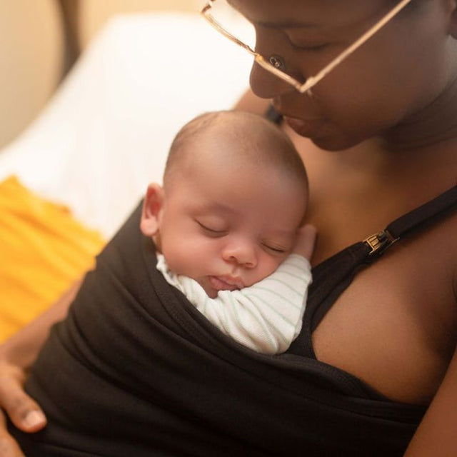 A baby is sleeping comfortably while being held skin-to-skin by their mother in a Joeyband.